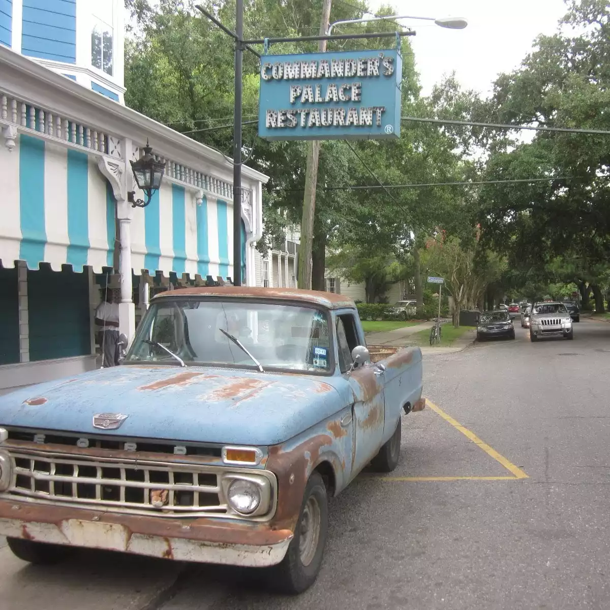 A vintage blue truck parked parallel in front of Commander's Palace in New Orleans. The tree-lined street stretches away in perspective.