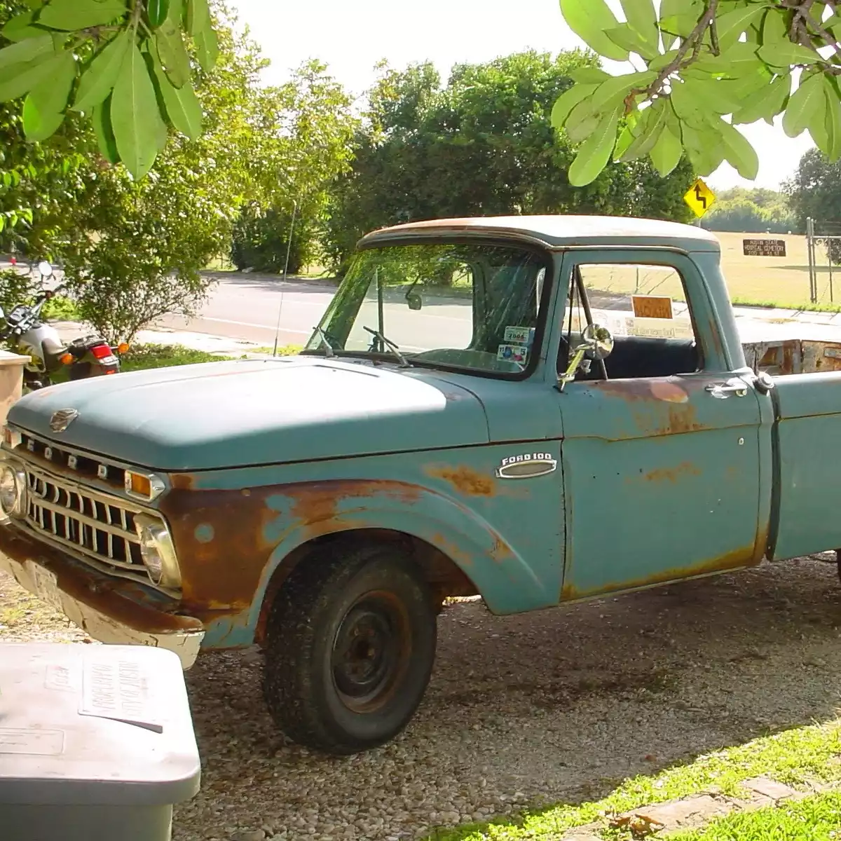 Oblique view of a vintage blue truck in a driveway with green lawn and leaves all around it.