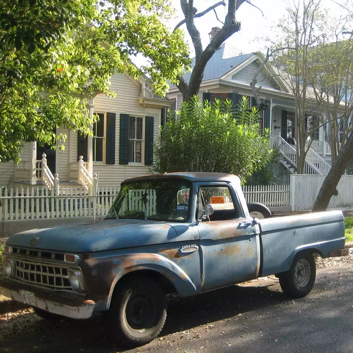 A vintage blue truck parked along the curb on a shady tree-lined avenue in front of a old-neighborhood house.