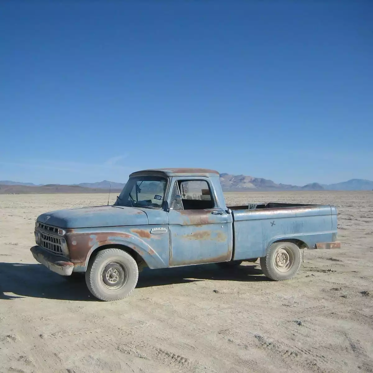 A vintage blue truck on the playa of Black Rock City in northwestern Nevada. The Painted mountains are distant. The tires of the truck are covered in grayish alkaline dust.