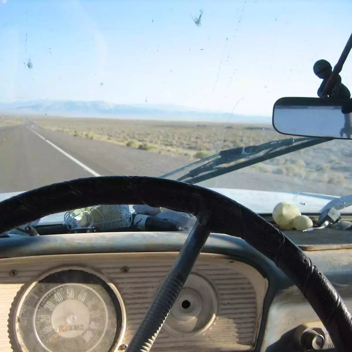 Point of view of driving an old truck on a two lane highway into vast scenery. A thin out-of-round steering wheel, dust-covered speedometer, strange objects on a dirty steel dashboard, comet shaped dead bugs, then the road stretching into a vanishing point under far-away mountains.